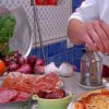 Un cuoco prepara una pizza circondato da salumi, verdure e spezie.