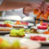 Mani di chef impiattano salmone e verdure su piatti bianchi.