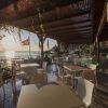 Tavoli bianchi con sedie sotto un pergolato si affacciano sul mare al tramonto.