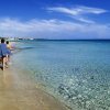 Due persone camminano su una spiaggia sabbiosa vicino al mare trasparente sotto un cielo azzurro.